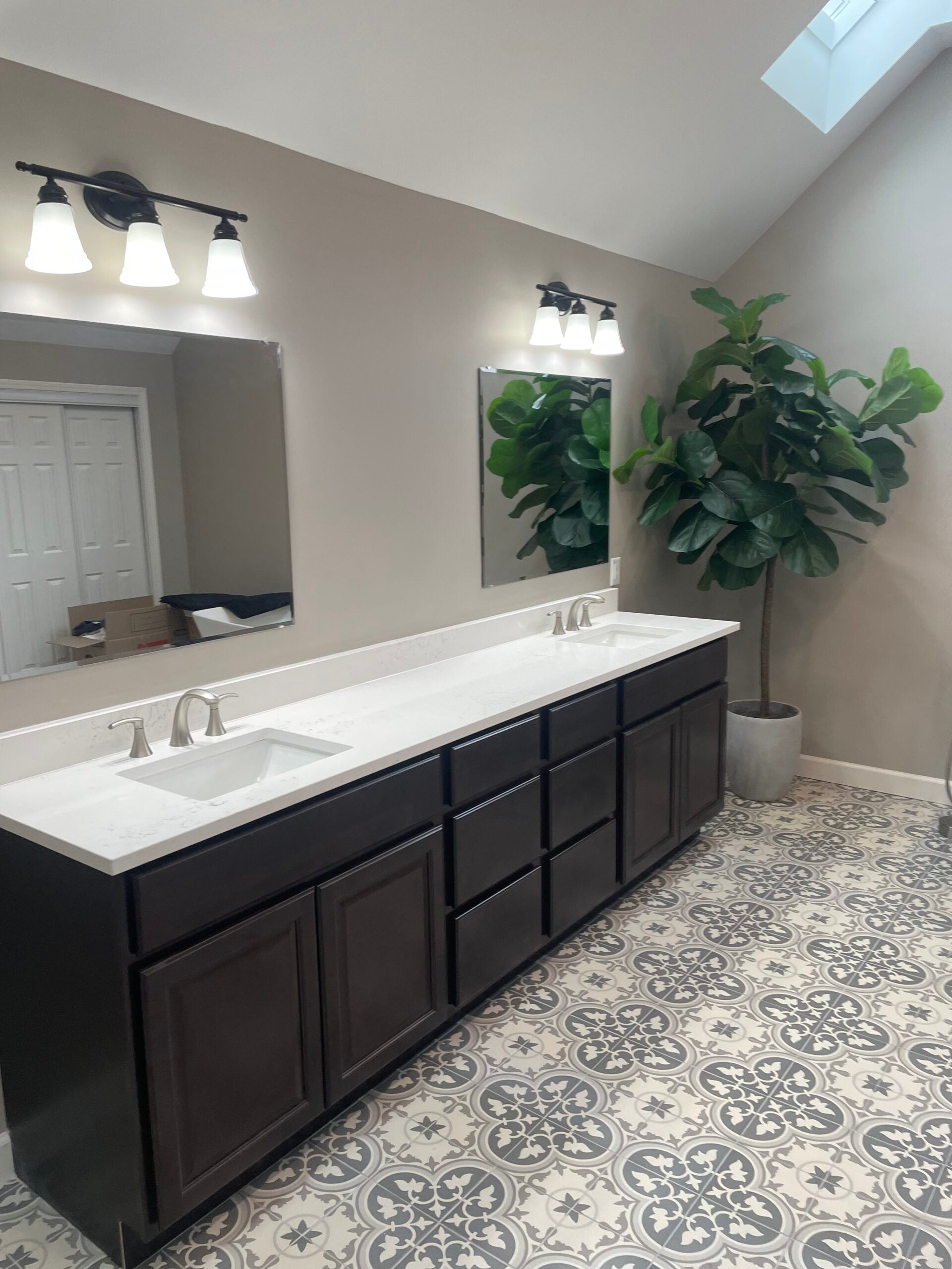 Remodeled bathroom with a long double vanity, white countertop, dark cabinetry, patterned tile flooring, and skylights.