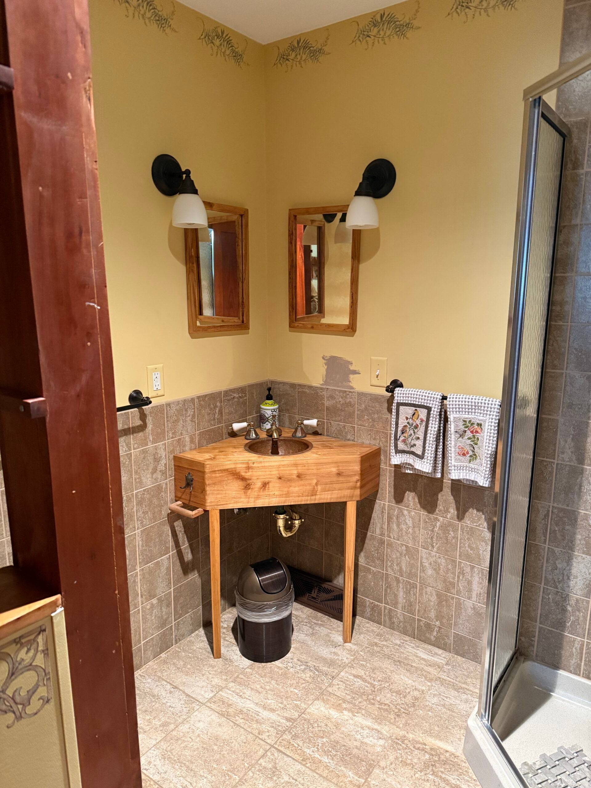 Bathroom before remodeling with an outdated corner vanity, dark tile, yellow walls, and enclosed shower.