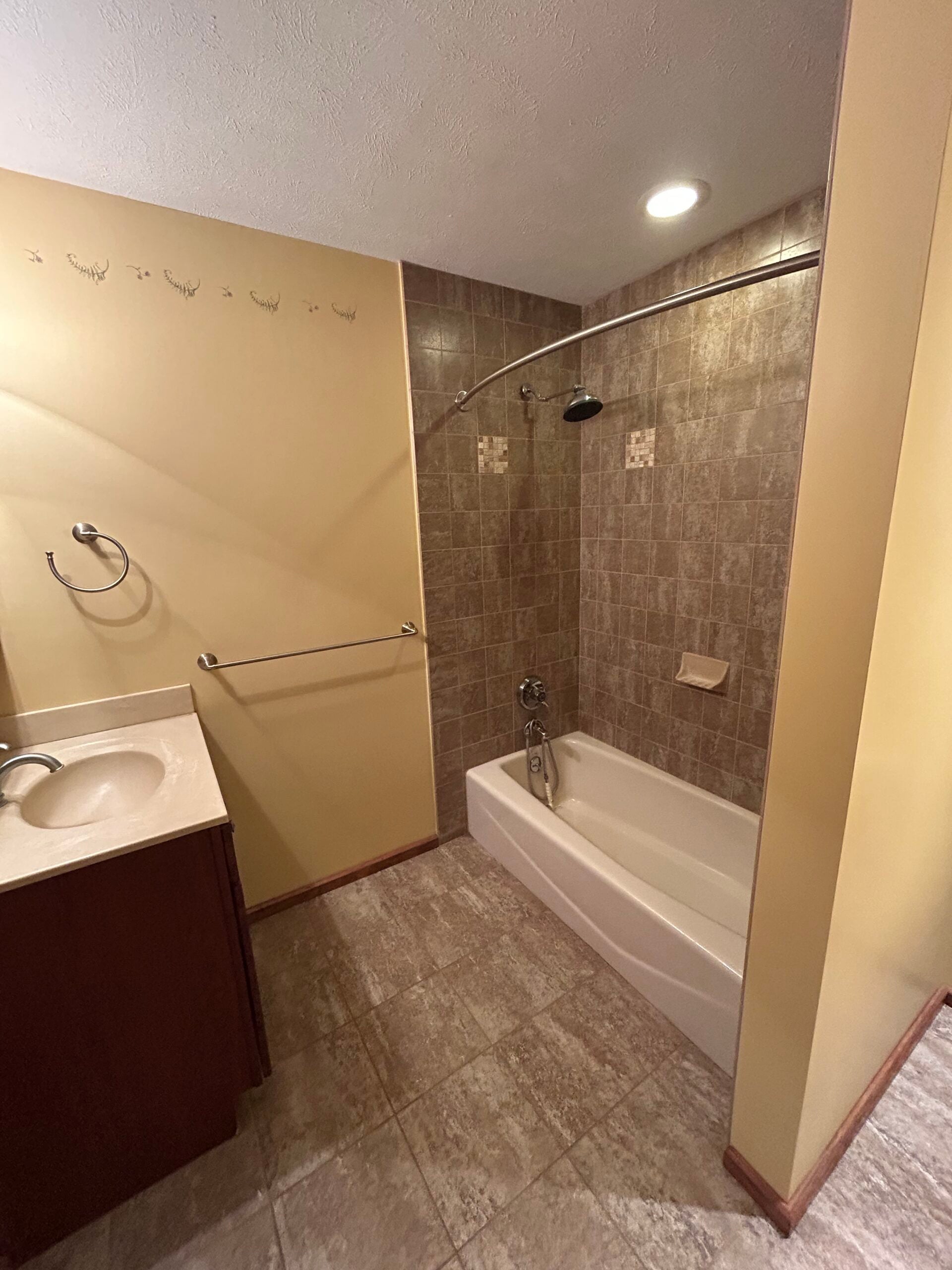 Bathroom before renovation with a tiled tub and shower surround, dated flooring, yellow walls, and traditional vanity.