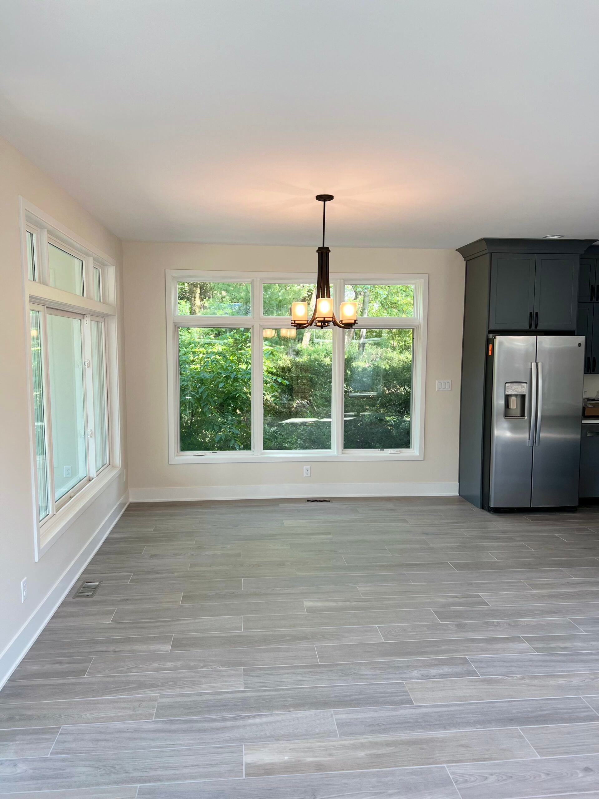 Open dining area with large windows, modern chandelier, gray flooring, and adjoining dark gray kitchen cabinetry.