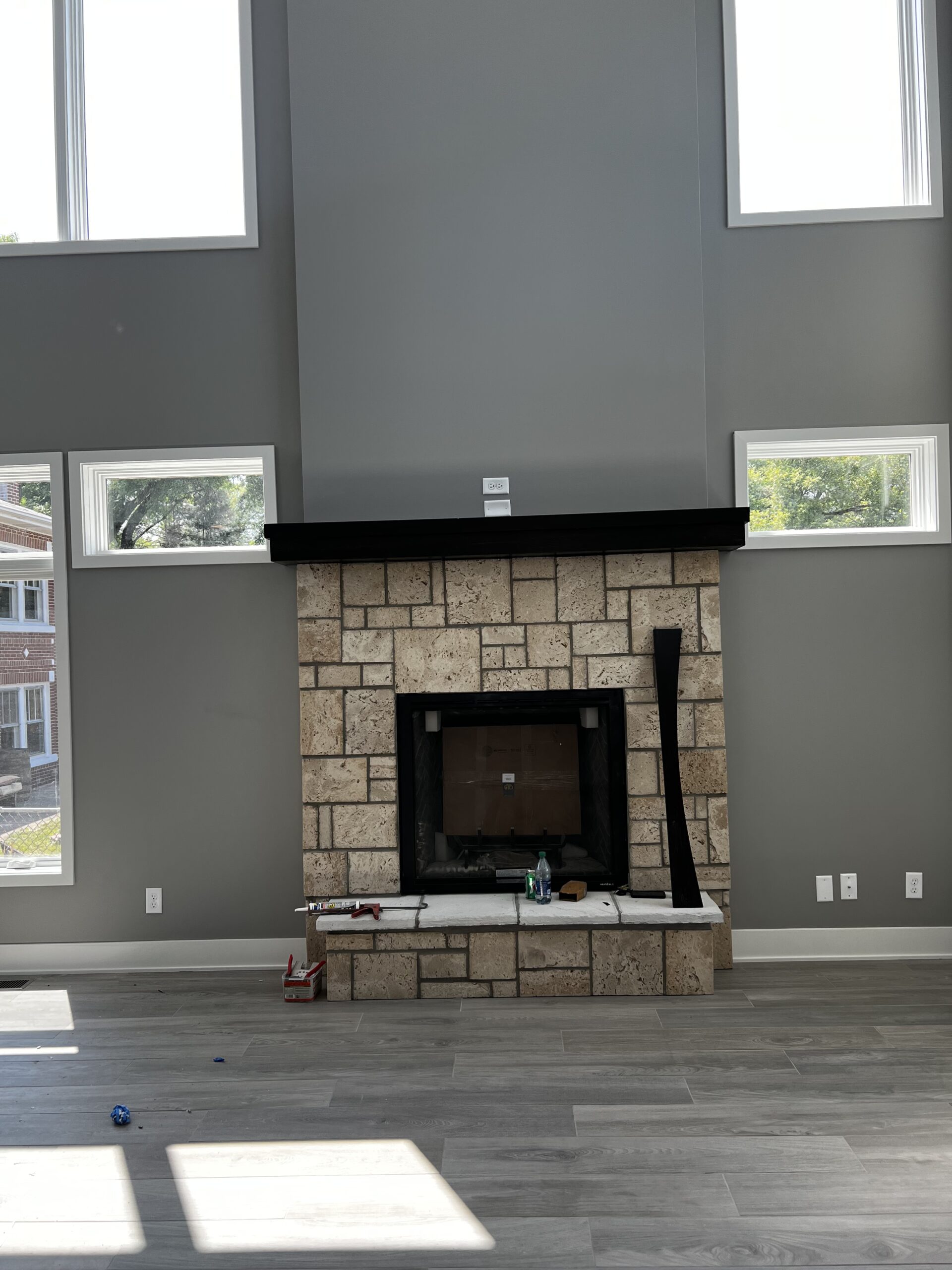 Custom stone fireplace with black mantel, tall gray accent wall, surrounding windows, and gray flooring.
