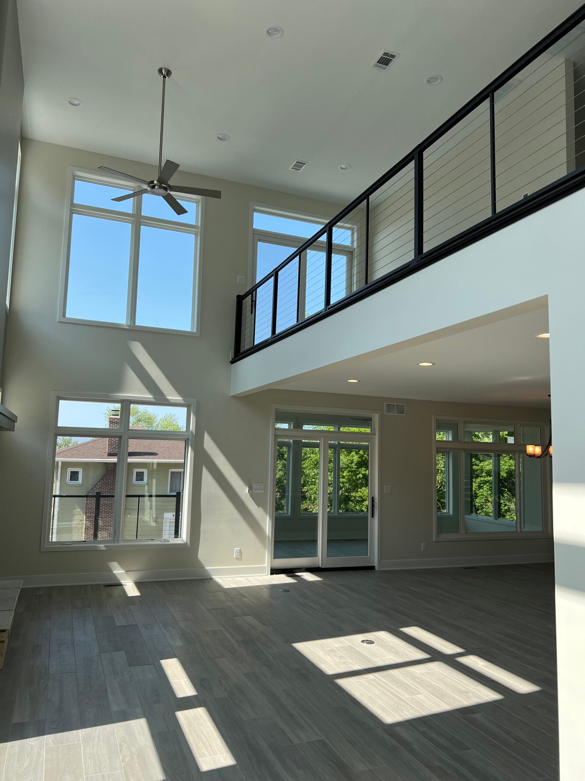 Bright two-story living room with large windows, gray flooring, and a black cable railing overlooking the main level.