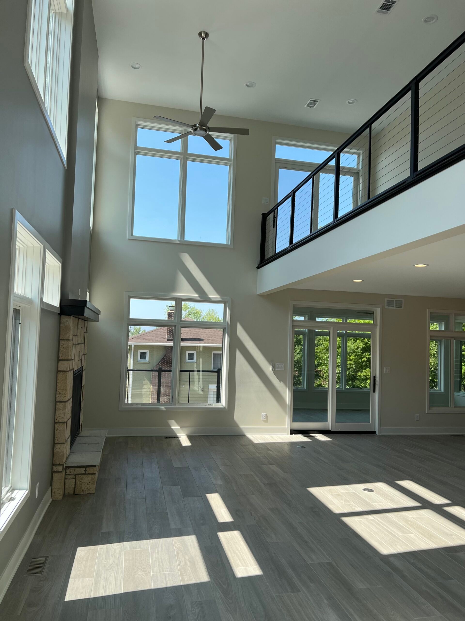 Bright two-story living room with large windows, gray flooring, stone fireplace, ceiling fan, and black cable railing.