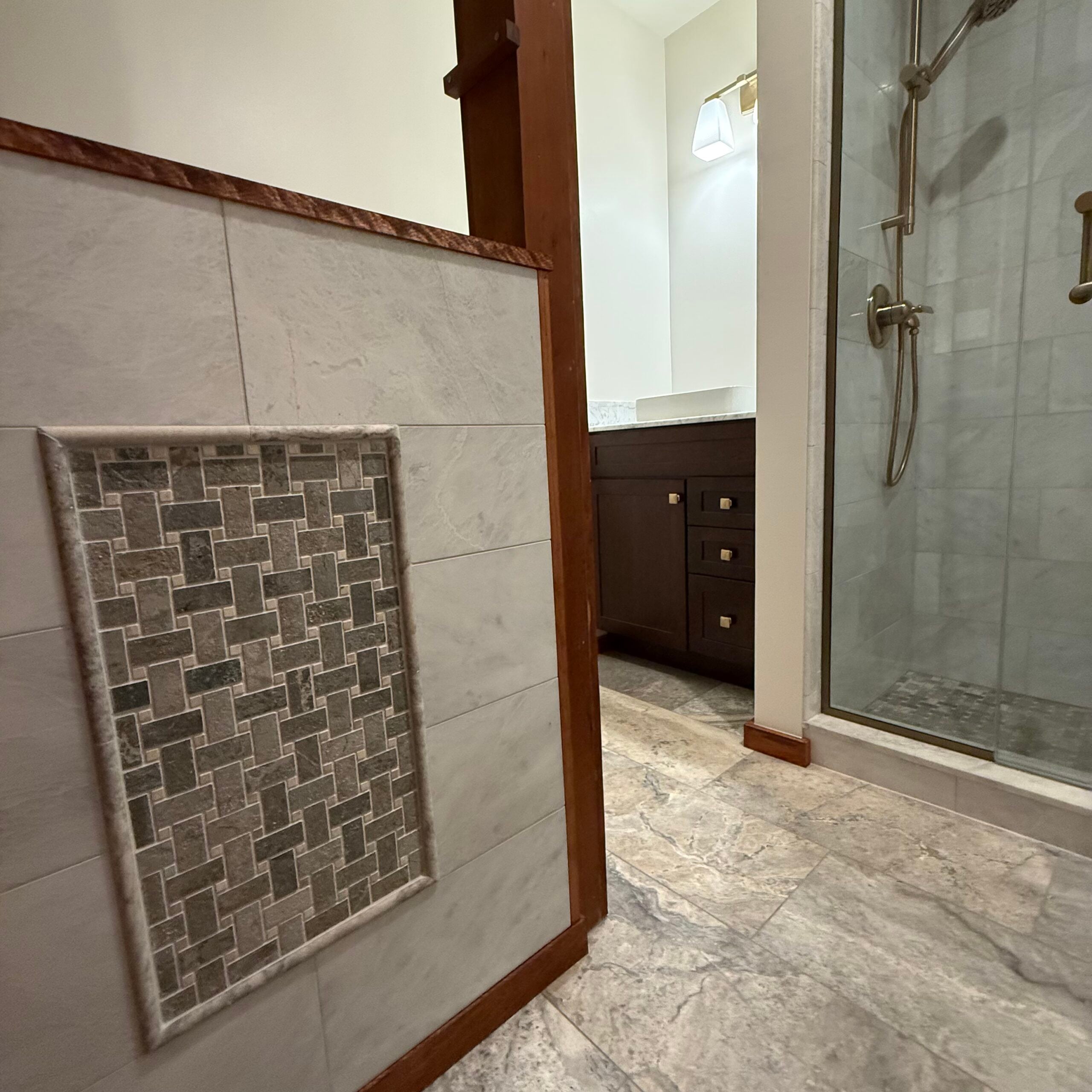 Remodeled bathroom featuring marble-look tile, a decorative mosaic wall inset, glass shower, and dark wood vanity.