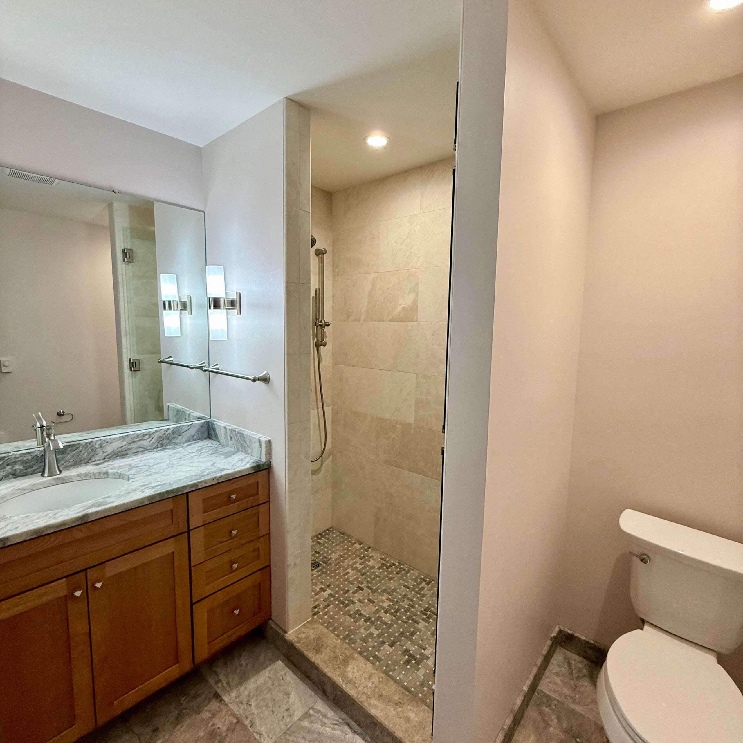 Remodeled bathroom with a wood vanity, stone countertop, walk-in tiled shower, mosaic floor, and separate toilet area.