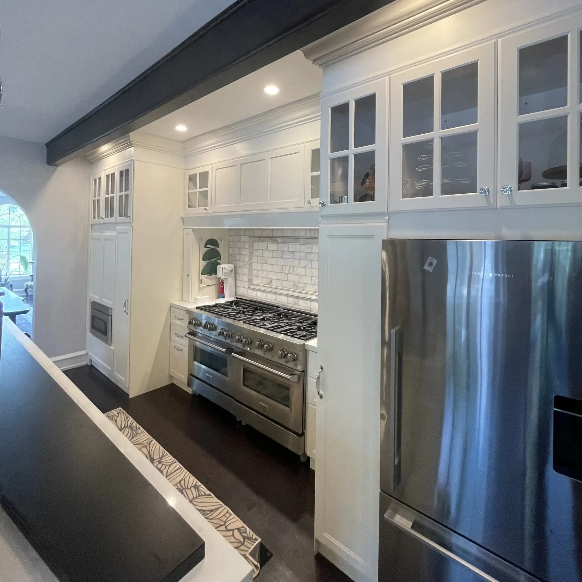 Custom kitchen remodel featuring white cabinetry, glass-front upper cabinets, stainless steel appliances, tile backsplash, and dark countertops.